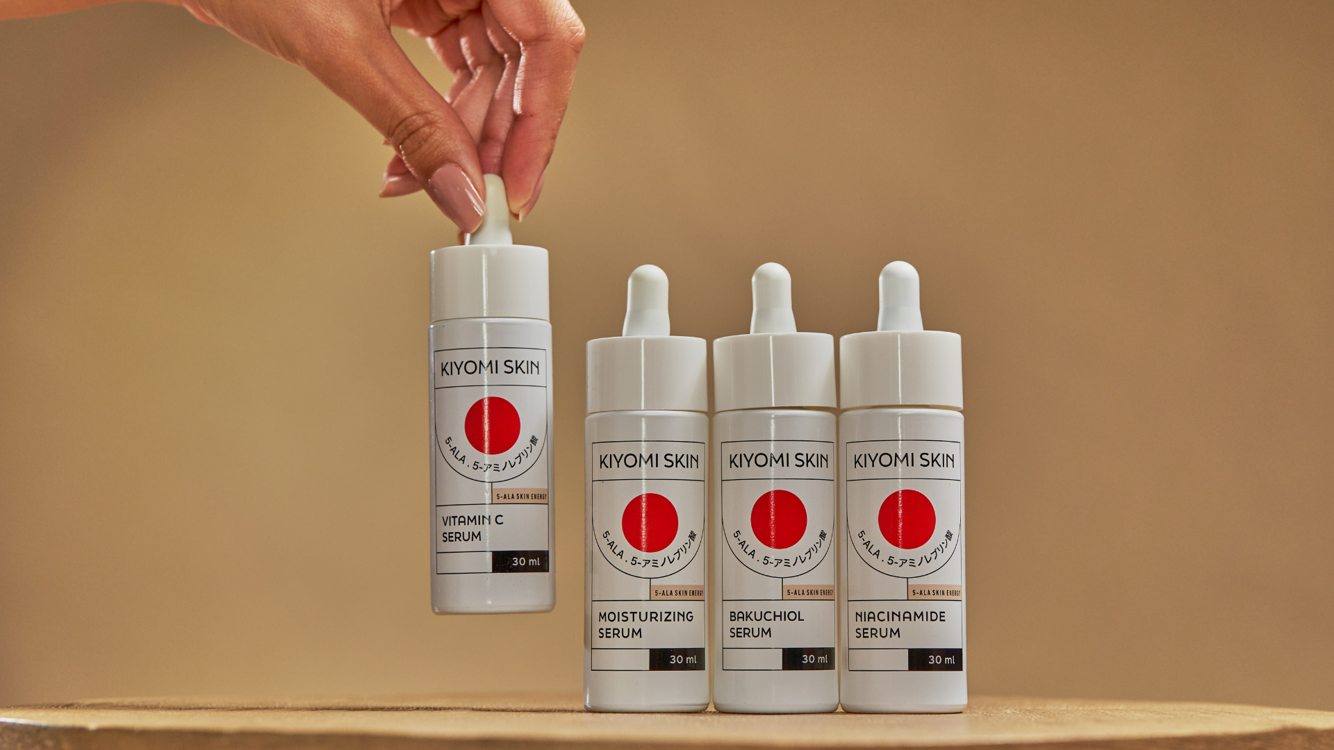 A hand lifts a Kiyomi Skin Vitamin C Serum bottle next to three other Kiyomi Skin serum bottles.