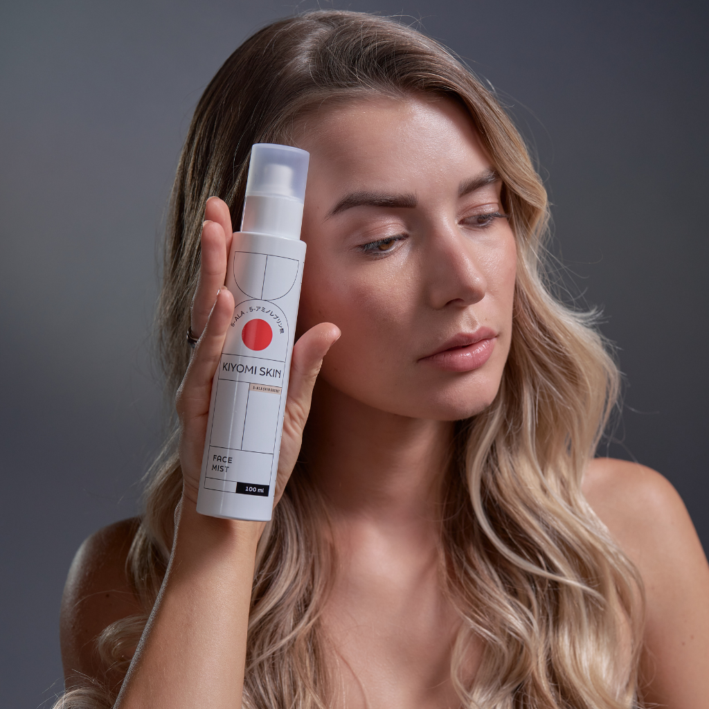 A woman with long blonde hair holds a Kiyomi Skin face mist bottle near her face.