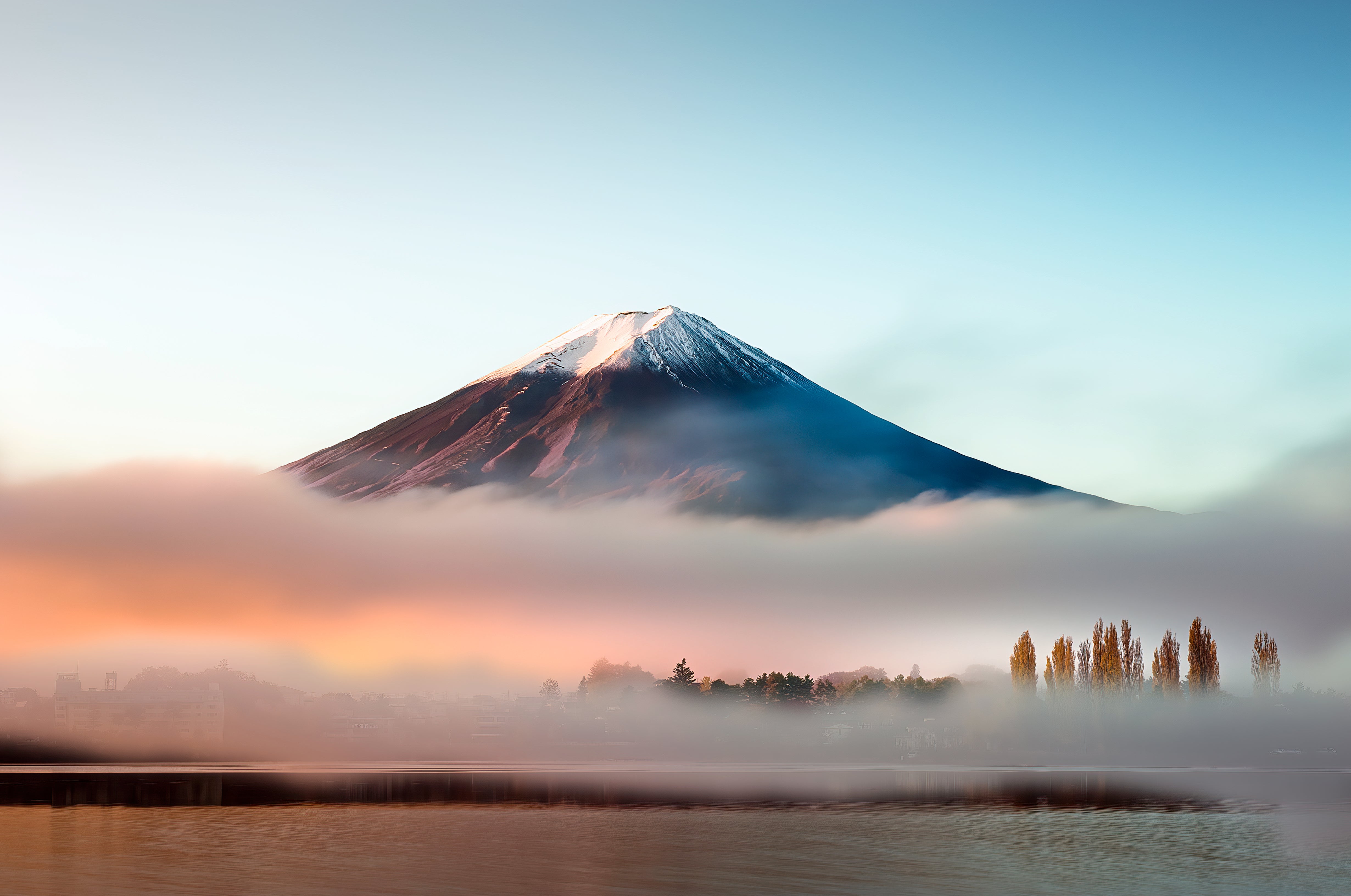 Snow-capped mountain with fog at sunrise, tall trees and water in the foreground.