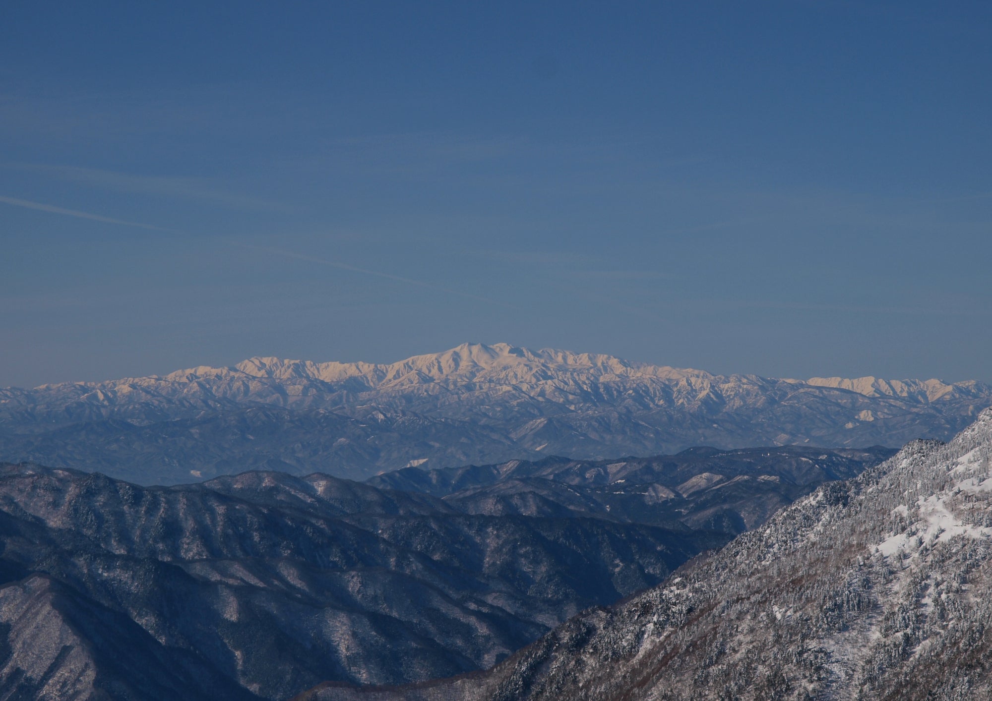 Snow-covered mountain range under a clear blue sky, with distant peaks illuminated by sunlight.