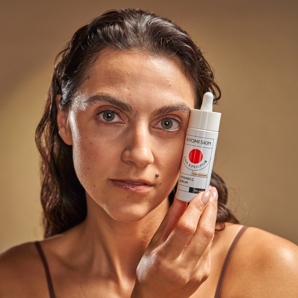 Woman with wet hair holds a Kiyomi Skin serum bottle next to her face, looking directly at the camera.