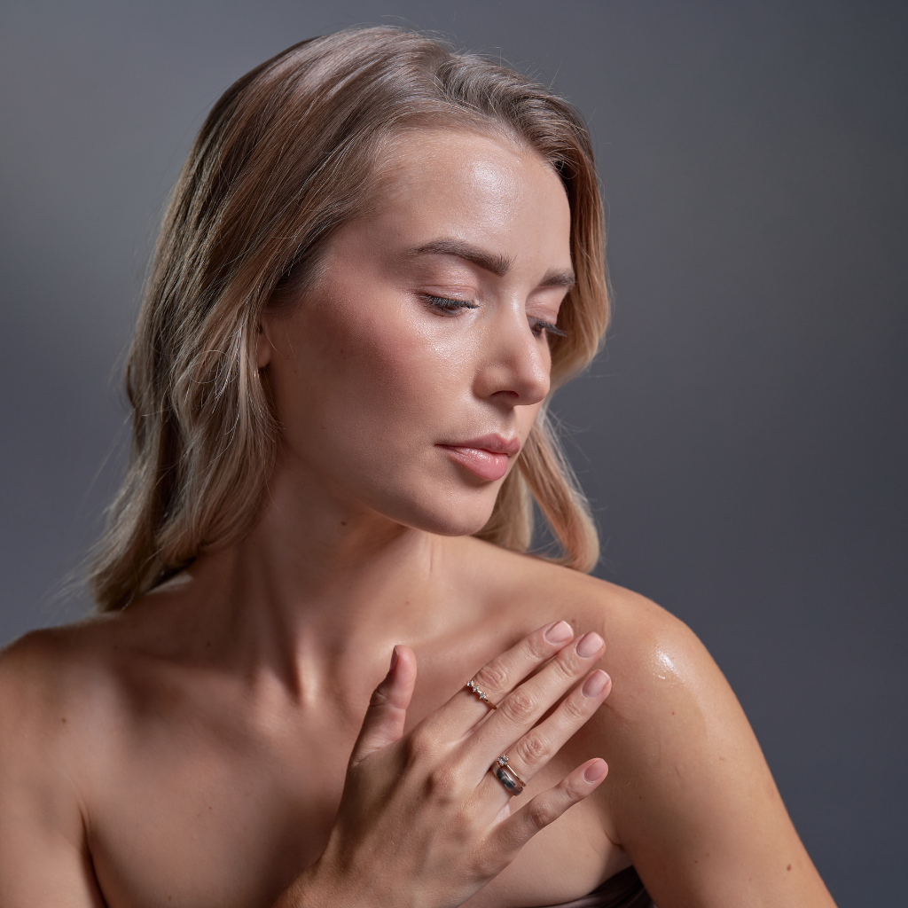 A woman with blonde hair touches her shoulder while looking down, against a neutral gray background.