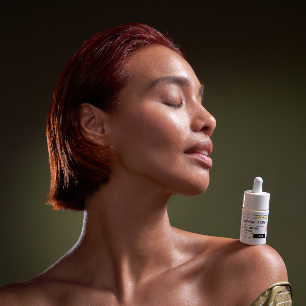 Woman with closed eyes poses with a skincare serum bottle balanced on her shoulder against a dark background.