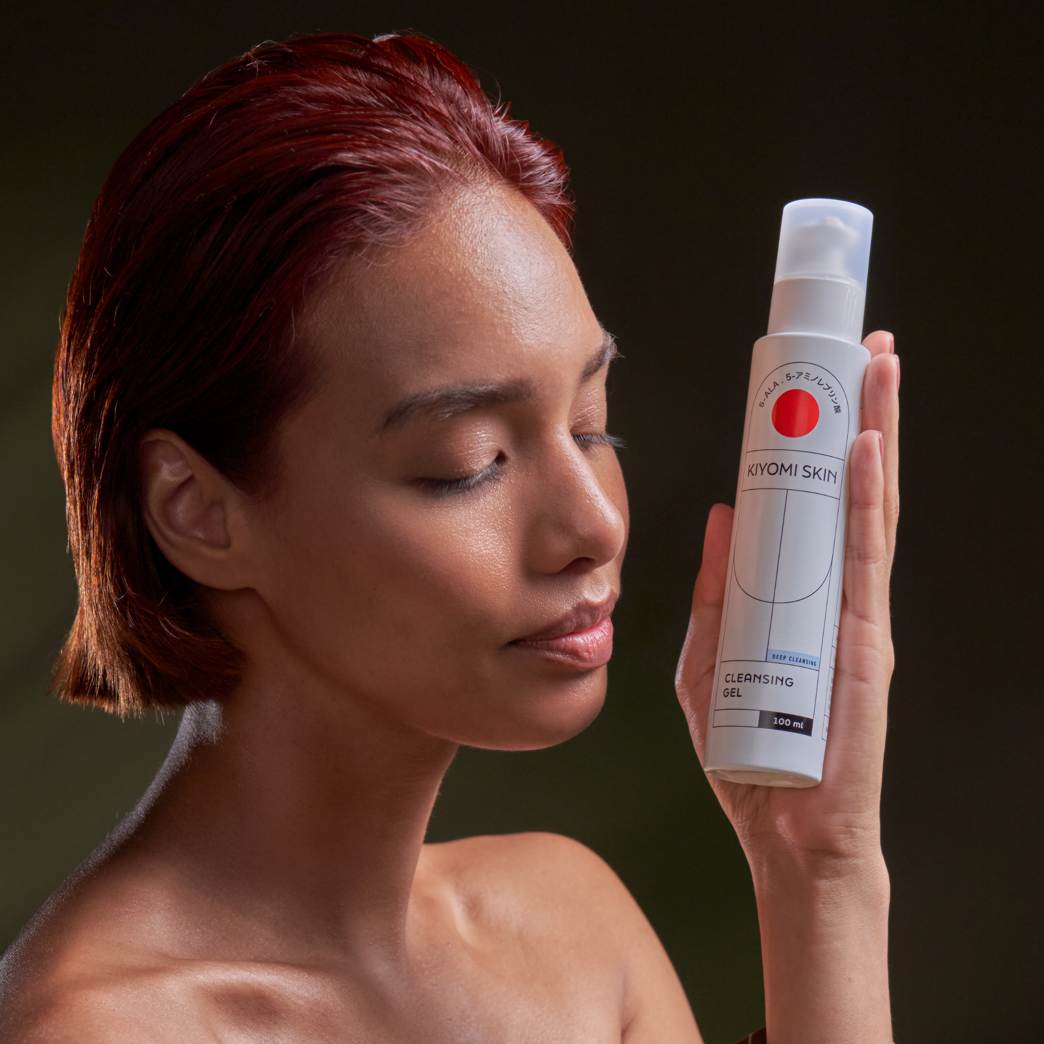 Red-haired woman holds a bottle of Cleansing Gel near her face, eyes closed, enjoying a moment of gentle facial cleansing.