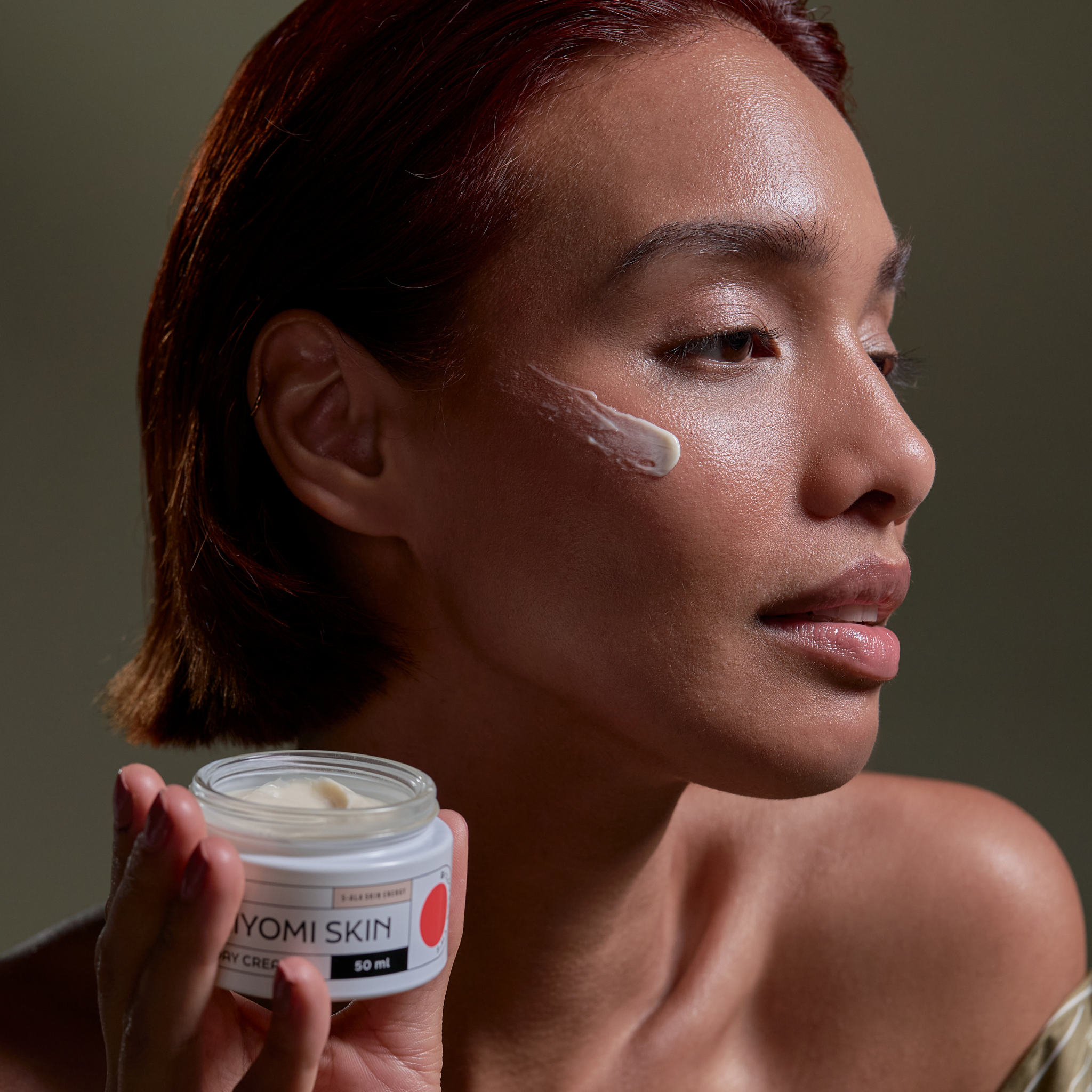 A woman applies Day Cream to her cheek while holding a 50 ml jar, a fragrance-free facial moisturizer and hydrating daily cream, perfect for gentle, effective skin care.