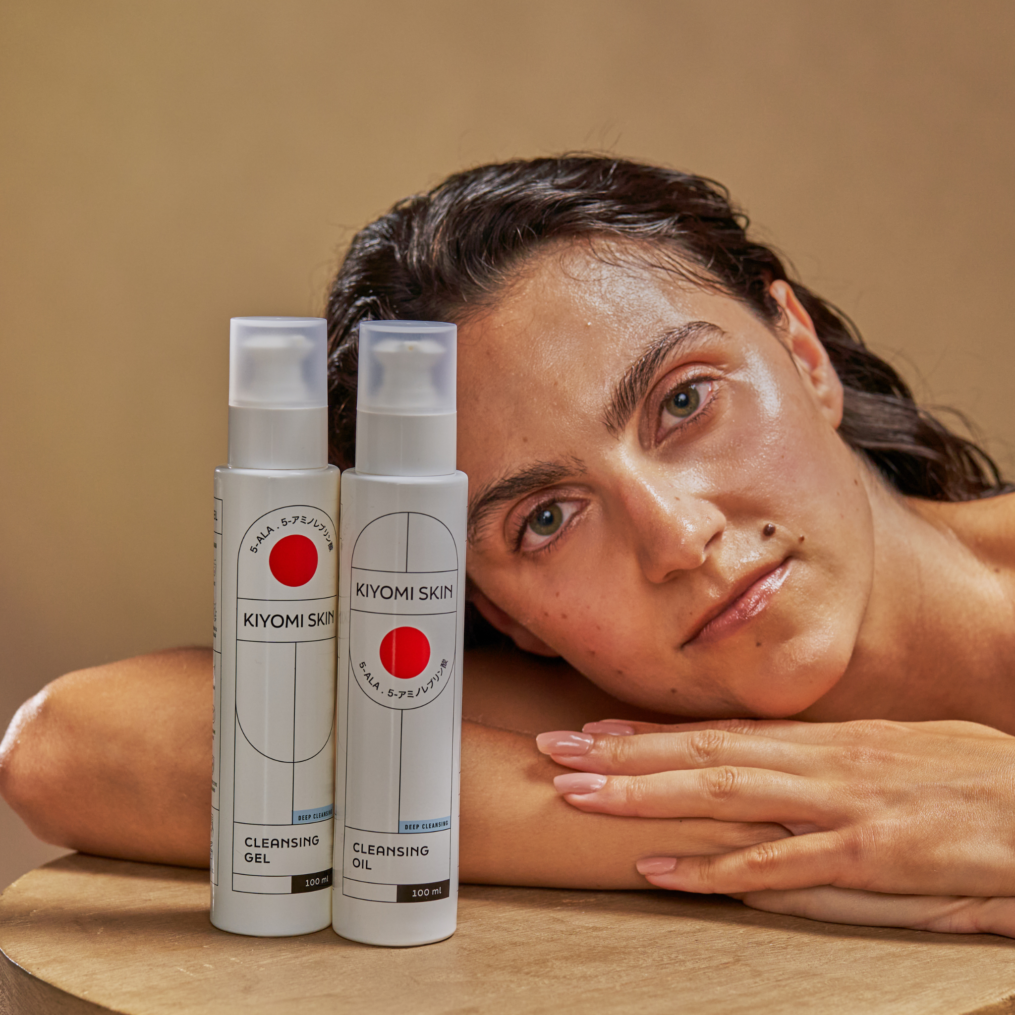 A woman rests her head on a table next to the Double Cleansing Set by Kiyomi Skin, highlighting the products against a neutral background for deep cleansing (porentiefe Reinigung).
