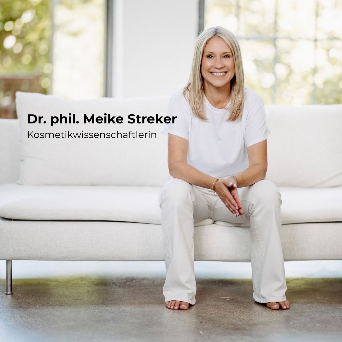 Eine Frau mit langen blonden Haaren in weißem Hemd und weißer Hose sitzt lächelnd auf einem Sofa. Text: Dr. phil. Meike Streker.
