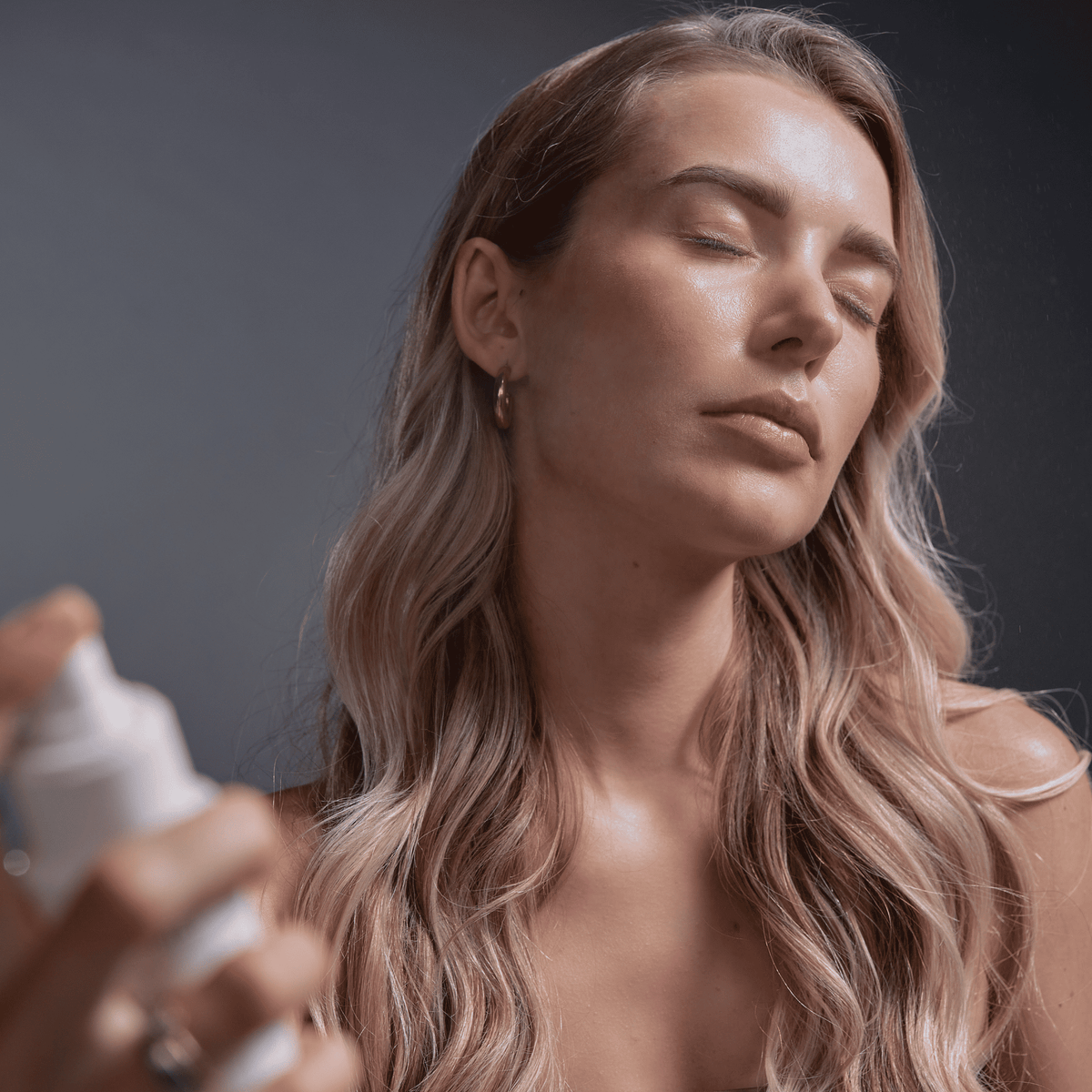 Eine Frau mit langen blonden Haaren sprüht sich vor einem dunklen Hintergrund mit geschlossenen Augen das „SBI ALApromo Face Mist“ mit Hyaluron ins Gesicht.