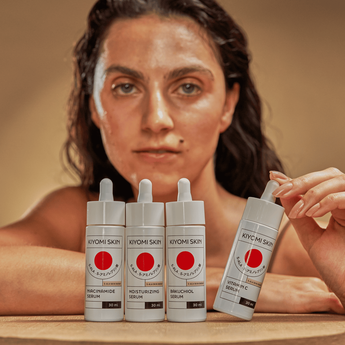 A woman poses with four Full Spectrum Serum Kit bottles, highlighting her skincare routine and the benefits of using a complete serum set displayed on the table in front of her.