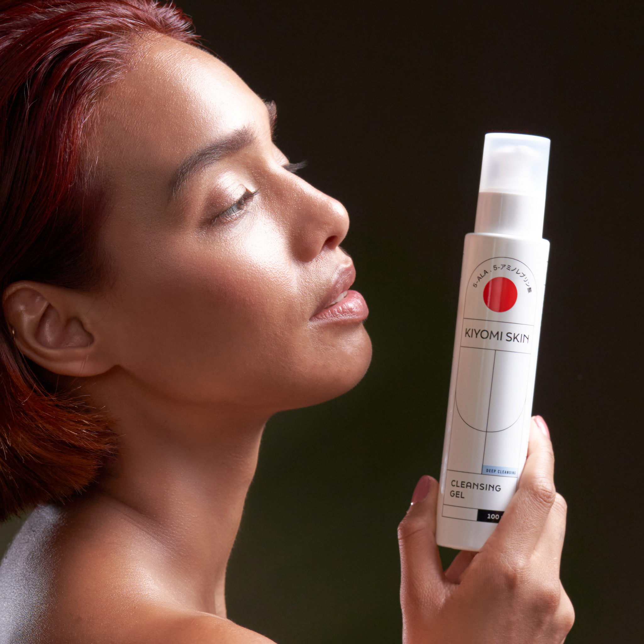 A woman with short red hair holds a Glass Skin Starter Duo bottle near her face against a dark background, highlighting the ideal addition to your skincare routine.