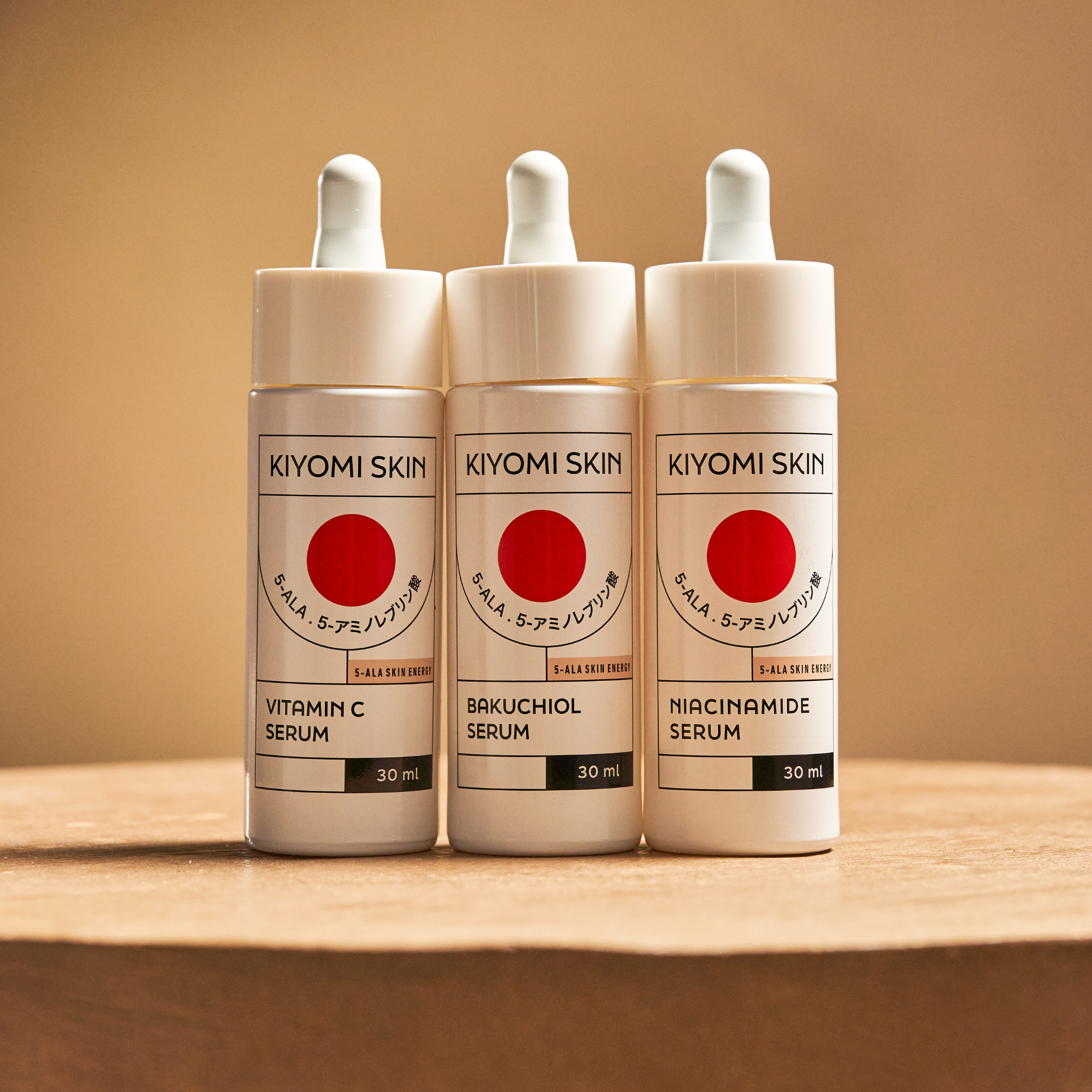 Three Kiyomi Skin serum bottles—Vitamin C, Bakuchiol, and Niacinamide—stand on a wooden surface, highlighting the Serum Trilogy Radiance & Renewal set for your skincare routine.