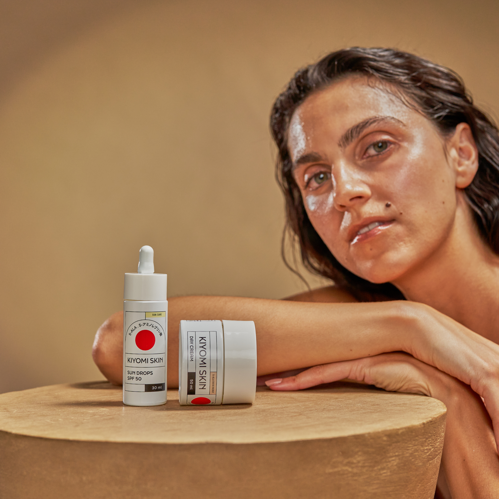 A woman with dewy skin poses with Skin Protection Duo on a round beige table against a neutral background.