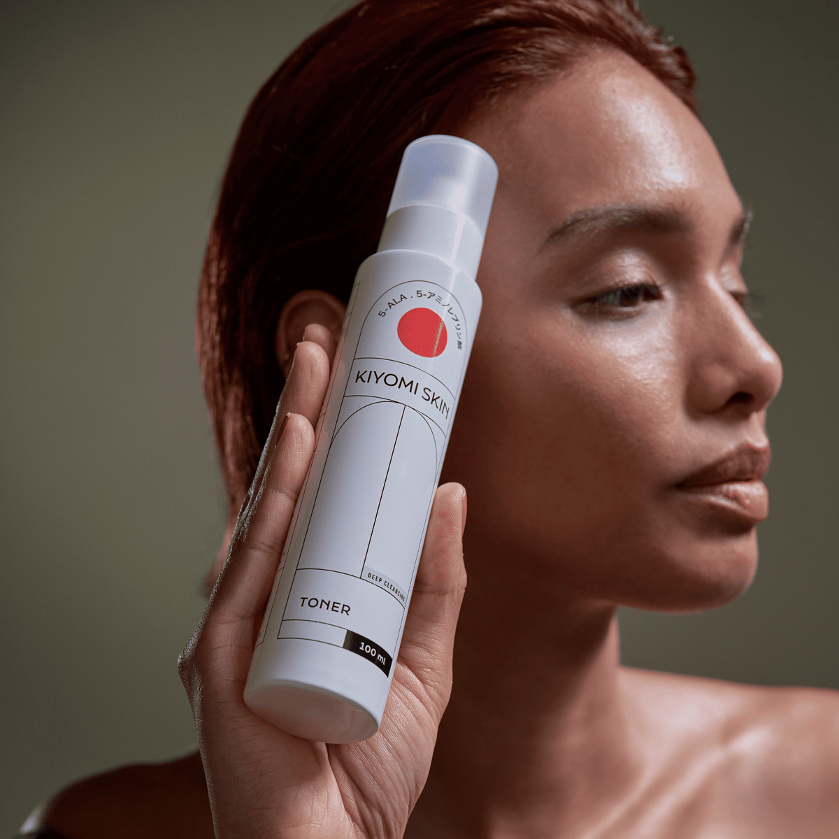 A woman holds a Toner bottle next to her face against a neutral background, highlighting her moisturizing routine.