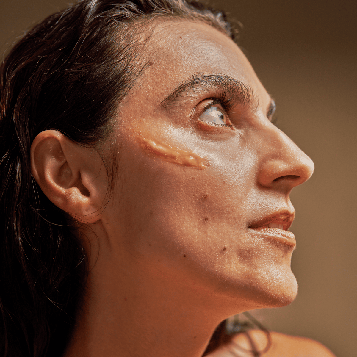 Woman with wet hair has a prominent scar on her cheek, looking upward, neutral background—her clear skin hints at the revitalizing effects of Vitamin C Serum.