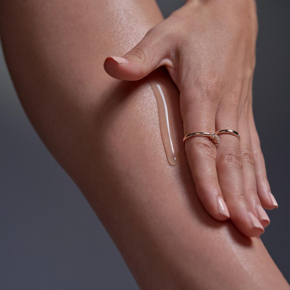 Eine Hand mit Ringen trägt transparentes Gel auf ein nacktes Bein vor einem neutralen Hintergrund auf.