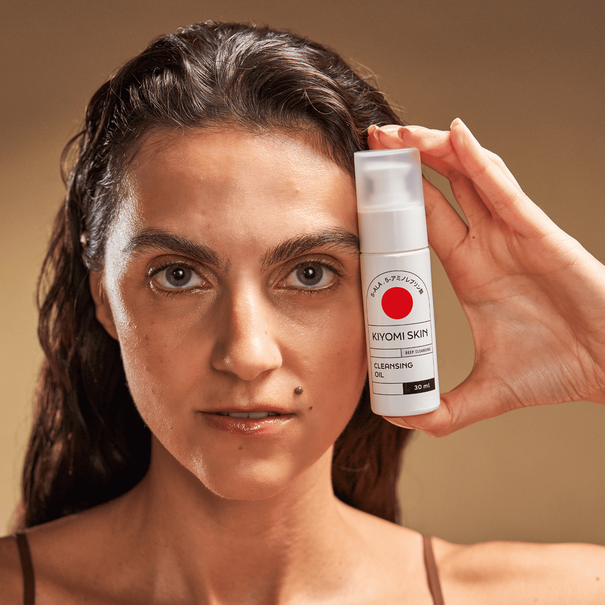 Eine Frau hält eine Flasche „SBI ALApromo Travel Size Cleansing Oil“ neben ihr Gesicht und blickt direkt in die Kamera – die sanfte Formel ist ideal für empfindliche Haut und in einer praktischen Reisegröße erhältlich.