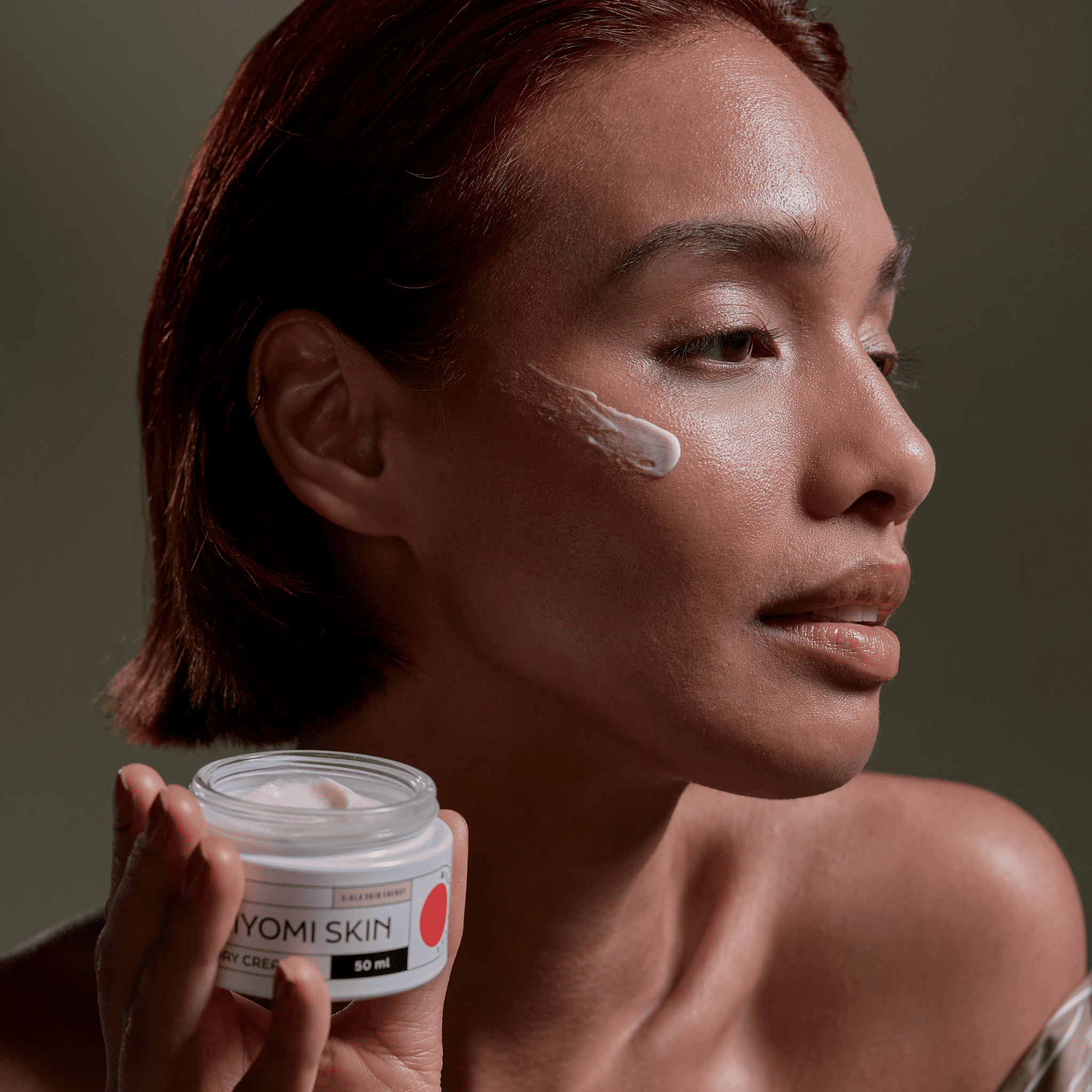 A woman applies Day Cream to her cheek while holding a 50 ml jar, a fragrance-free facial moisturizer and hydrating daily cream, perfect for gentle, effective skin care.
