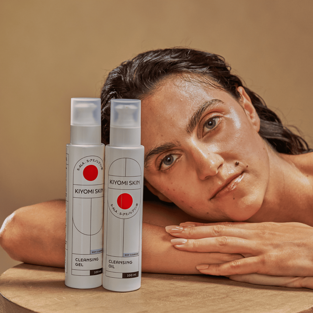 Eine Frau legt ihren Kopf auf den Tisch neben zwei Produkten aus dem „SBI ALApromo Double Cleansing Set“, darunter ein Reinigungsöl, vor einem neutralen Hintergrund.