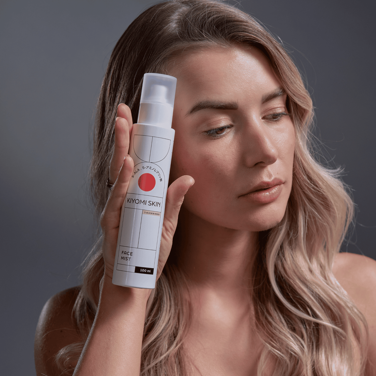 Eine Frau mit langen Haaren hält vor einem neutralen Hintergrund eine Flasche „SBI ALApromo Face Mist“ nahe an ihr Gesicht und präsentiert damit die feuchtigkeitsspendende Hyaluronsäure-Formel des Produkts.