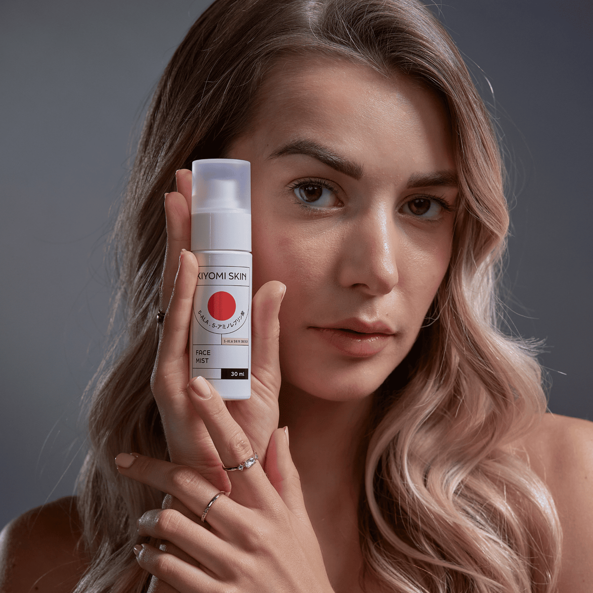 Eine Frau mit langen blonden Haaren hält eine Flasche „SBI ALApromo Travel Size Face Mist“ nahe an ihr Gesicht und blickt in die Kamera. Dieses feuchtigkeitsspendende Gesichtsspray eignet sich ideal für die feuchtigkeitsspendende Gesichtspflege unterwegs.