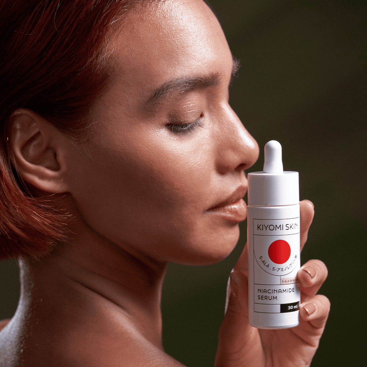 Eine Frau mit glatter, glasartiger Haut, die eine Flasche aus dem „Kiyomi Skin Glass Skin Starter Duo“ nahe an ihr Gesicht hält.