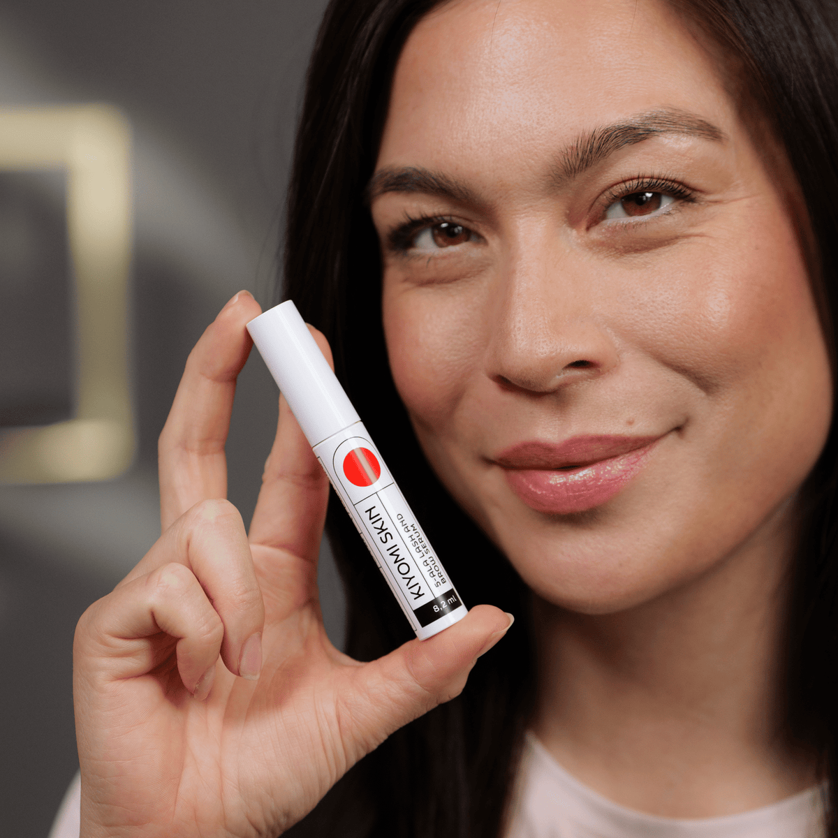Eine lächelnde Frau, die vor einem unscharfen Hintergrund eine Tube „Kiyomi Skin Lash and Brow Serum“ in der Hand hält.