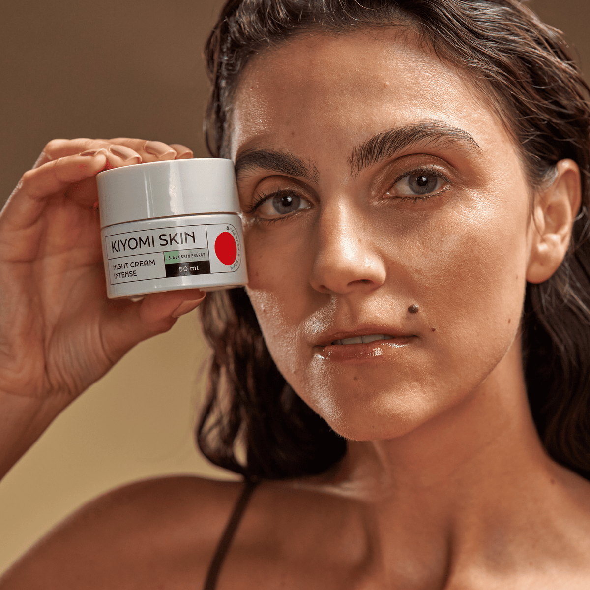 Eine Frau hält ein Töpfchen der „SBI ALApromo Night Cream Intense“ vor ihr Gesicht, vor einem neutralen Hintergrund.