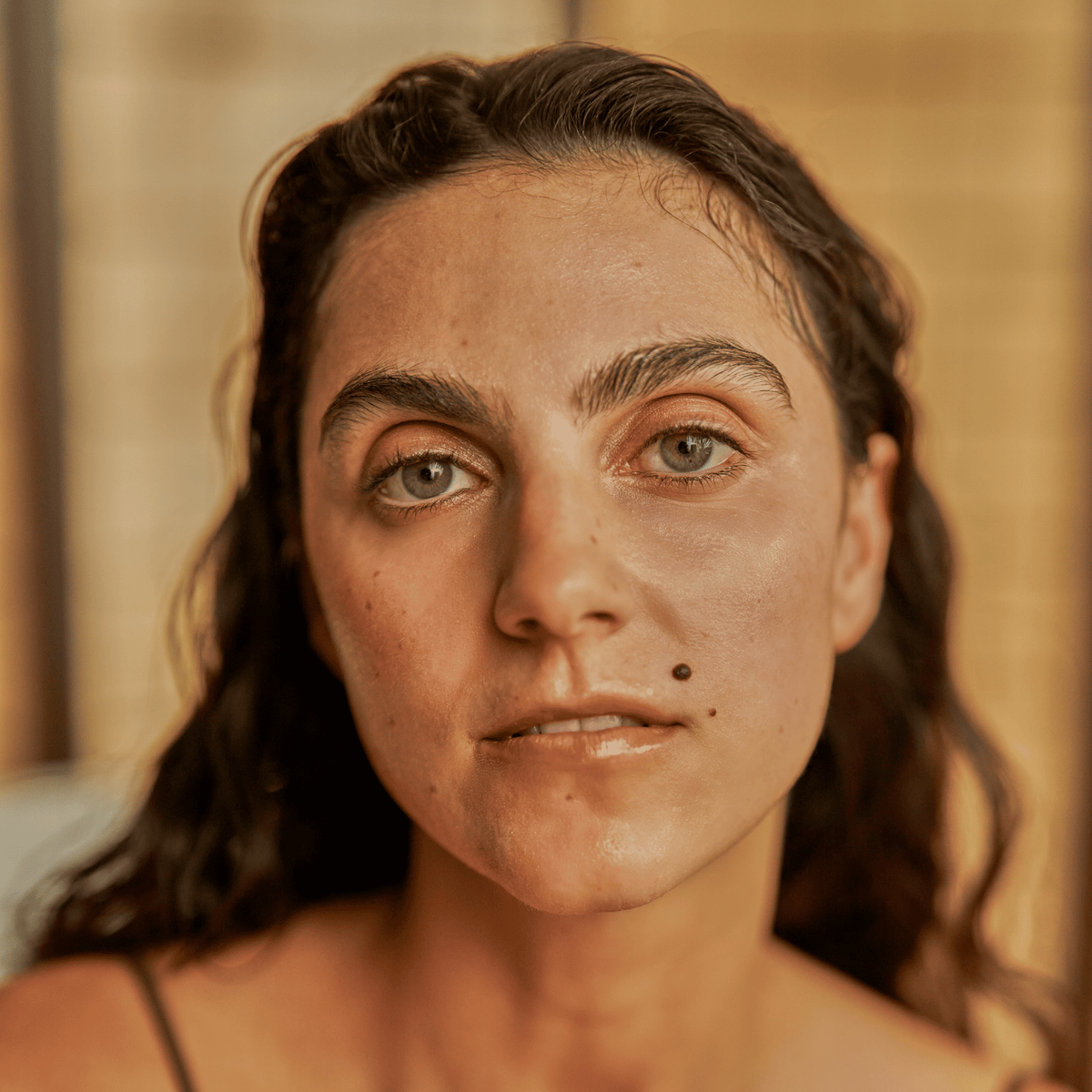 Woman with wavy dark hair and a prominent mole looks into the camera, softly lit against a neutral background—her complexion appears radiant and hydrated, as if refreshed by the Sheet Mask.