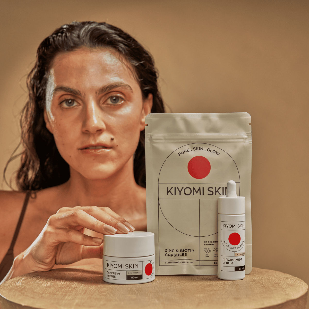 Eine Frau posiert vor dem „Skin Glow Essentials – Inner and Outer Glow“-Set von SBI ALApromo, das eine Tagescreme, ein Serum und eine Tüte mit mikronährstoffreichen Kapseln auf braunem Hintergrund enthält – perfekt für die ganzheitliche Hautpflege.