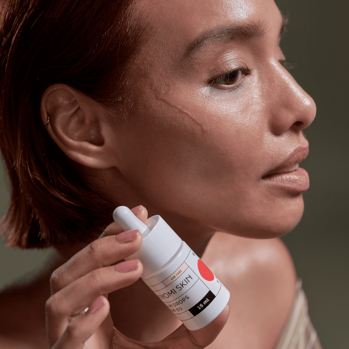 Eine Frau mit einer Narbe im Gesicht hält eine kleine Flasche „SBI ALApromo Travel Size Sun Drops SPF 50“ dicht an ihre Wange und unterstreicht damit den sanften Schutz und den optimalen UV-Schutz für einen leichten Sonnenschutz.